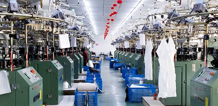 Wide panoramic shot of China seamless apparel manufacturing workshop, full row of automatic circular seamless knitting looms, finished white bodysuit blanks hanging on production line, plastic material storage bins, professional factory workers, industrial mass production setup for custom sports bra, shapewear, yoga wear OEM & ODM bulk manufacturing.