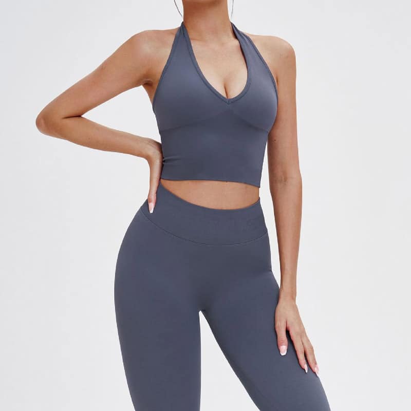 Side studio shot of womenโs dark gray seamless workout set, deep V halter racerback cropped sports bra with medium support, matching high rib waist full-length leggings, buttery naked-feel nylon spandex fabric, squat-proof stretch knit for yoga, gym and daily athleisure, OEM halter racerback sports bra legging set wholesale manufacturer.