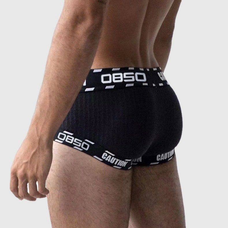 Rear close-up studio shot of menโs black seamless trunk underwear, textured vertical rib knit fabric, wide jacquard elastic waist & leg bands printed with custom brand text "OBSO CAUTION", 4-way stretch breathable knit, sweat-wick performance material for daily and athletic wear, OEM private label menโs intimates factory supply.